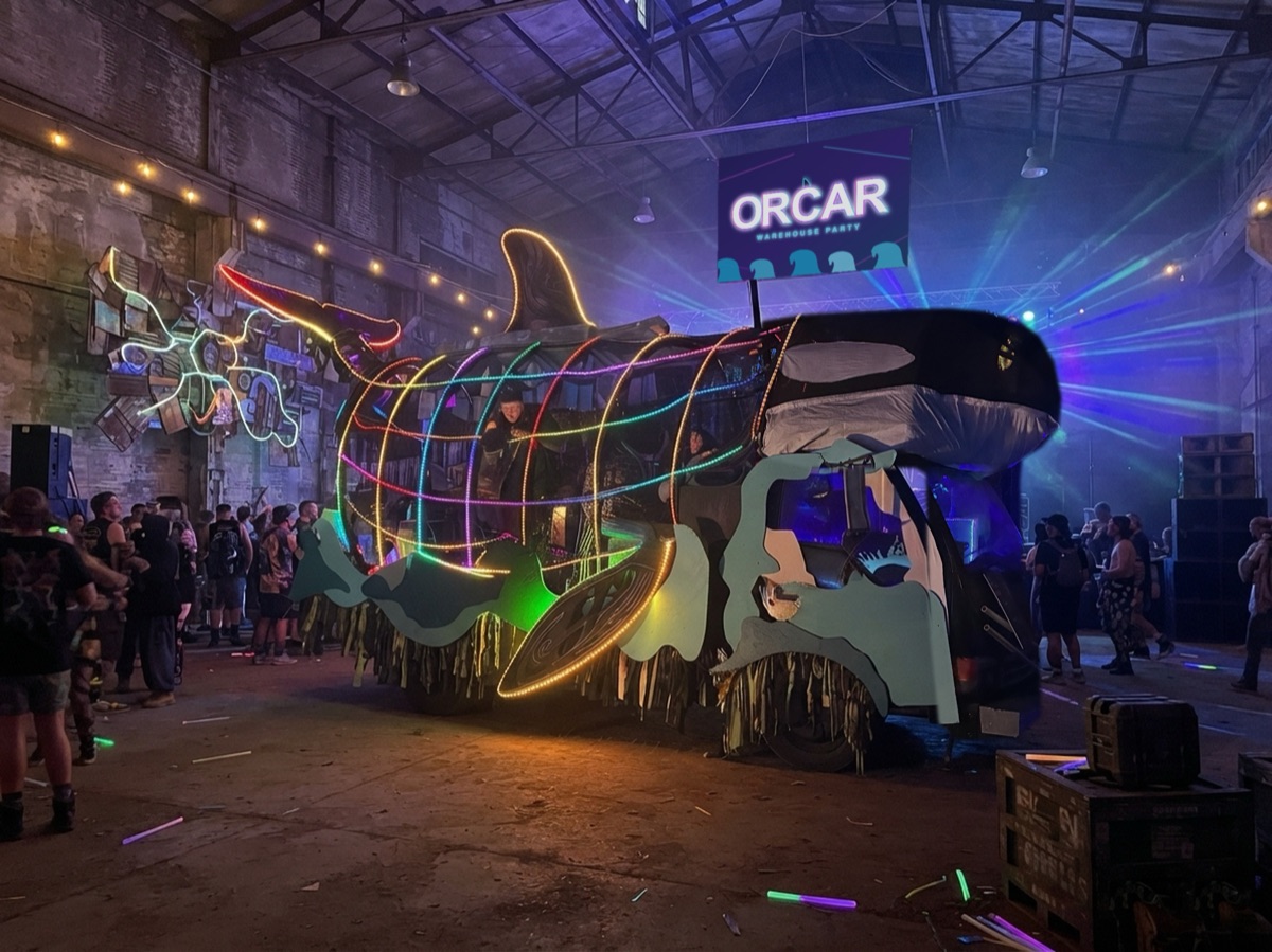 Illustration: the Orcar lit in rainbow LEDs inside a warehouse party, with a crowd and an Orcar Warehouse Party sign